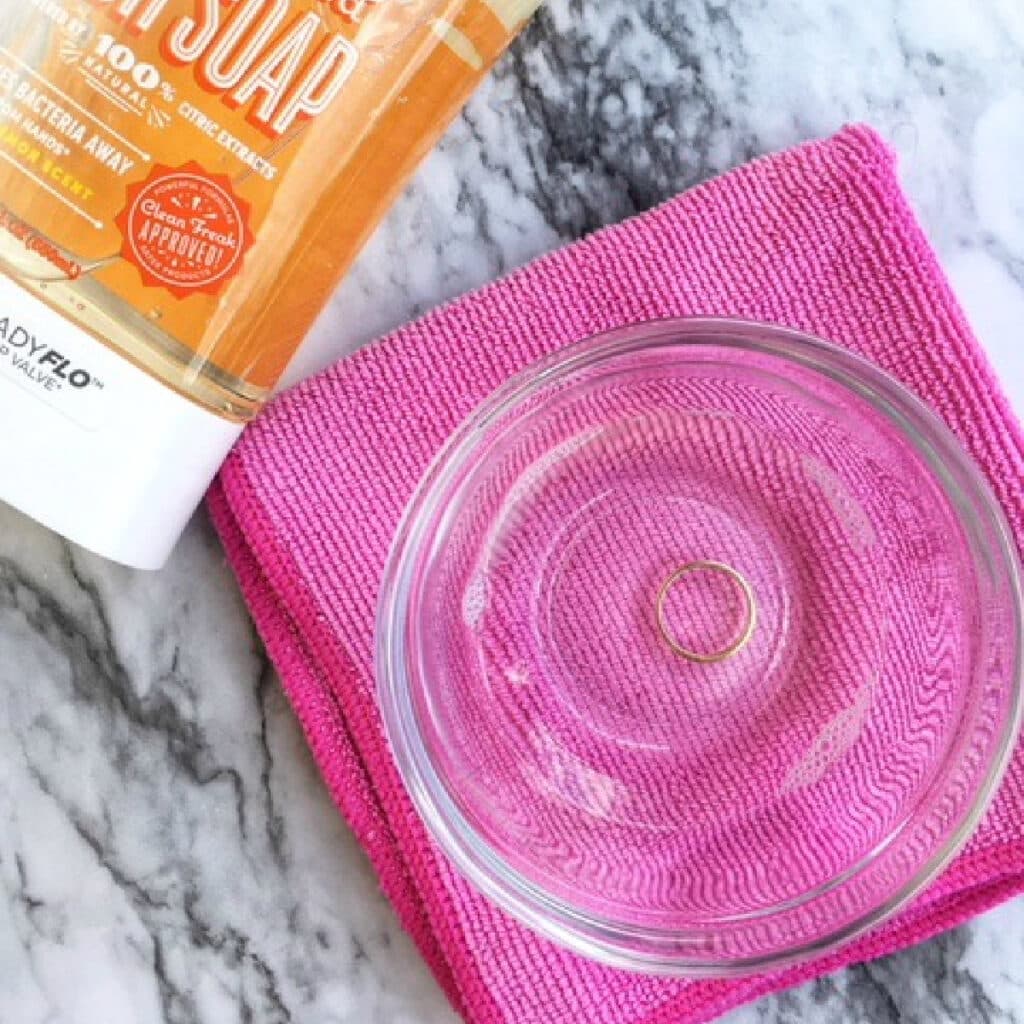 gold ring in a glass bowl of diy jewelry cleaner on top of a microfiber cloth