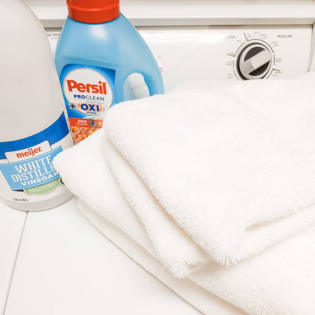 Towels, laundry detergent, and white vinegar on the washing machine before cleaning a plastic shower curtain.